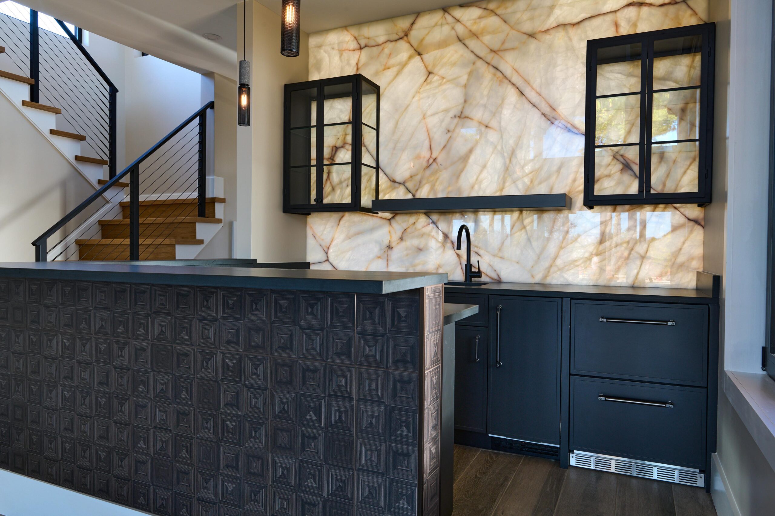 modern home bar featuring a backlit onyx wall, dark cabinetry, textured bar front, and a staircase in the background.